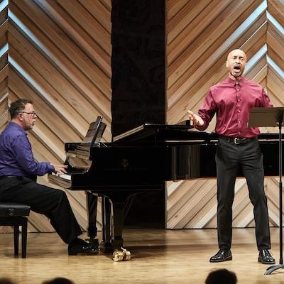 Action shot of pianist Jeffrey Sykes and bass-baritone Timothy Jones performing on stage. On the left, Jeffrey plays a black Steinway grand piano in a purple shirt. On the right, Timothy stands with arms outstretched, singing with an expressive, open-mouthed expression in a maroon button-down shirt and black trousers. They are positioned in front of a geometric wood-paneled backdrop.